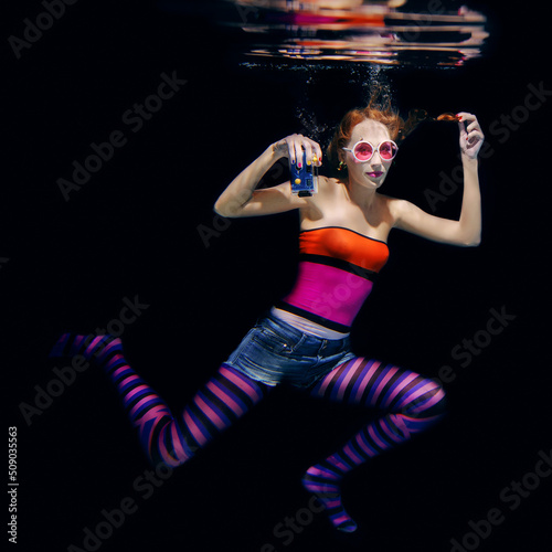 redhead funny woman in colorful clothes and pink sunglasses on the dark background swimming underwater with photo camera. Travel, sightseeing concept © k8most