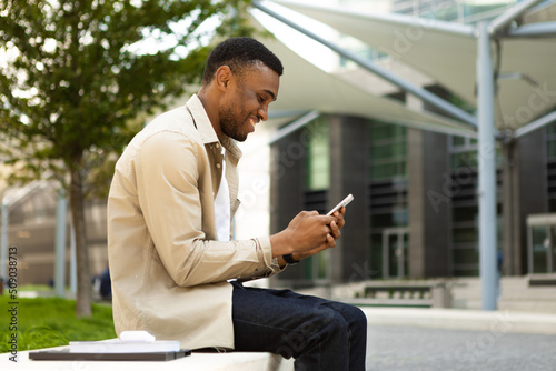 Side view of african american man using smart phone sitting on bench in city