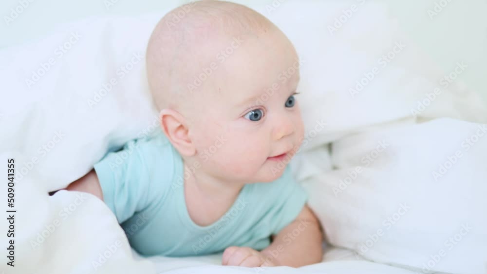funny caucasian baby infant lying in white bed on stomach in the morning, happy 3 month child in blue clothes awake in sunny day bedroom, smiling portrait of blue-eyed kid in white bedsheets funny caucasian baby infant lying in white bed on stomach in the morning, happy 3 month child in blue clothes awake in sunny day bedroom, smiling portrait of blue-eyed kid in white bedsheets