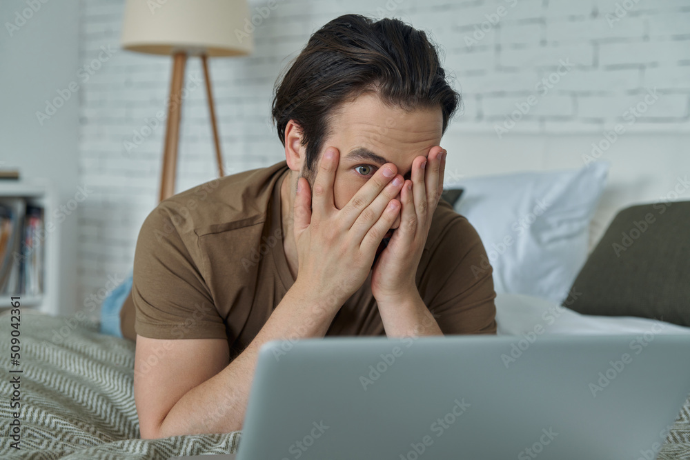 Terrified young man lying in bed and covering face with hand while looking at laptop Terrified young man lying in bed and covering face with hand while looking at laptop