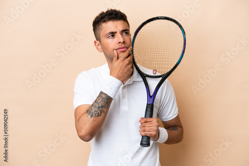 Young brazilian handsome man playing tennis isolated on beige background having doubts © luismolinero