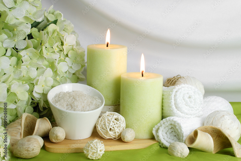 spa concept of soft green orchid flower with burning candles surrounded in natural dried elements, white towels, bath salts against a  white elegant swooped background. Spa still life