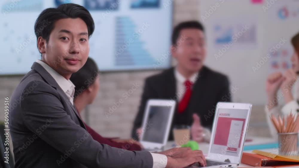 Confident young businessman sitting smiling at the camera in a meeting room with colleagues in the background. Confident young businessman sitting smiling at the camera in a meeting room with colleagues in the background.