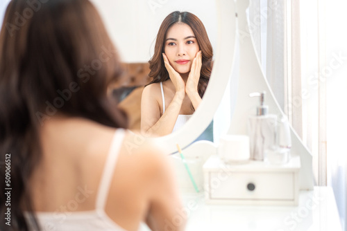 Smiling of young beautiful pretty asian woman clean fresh healthy white skin looking at mirror.asian girl touching on her face with hand and applying cream at home.spa and beauty concept