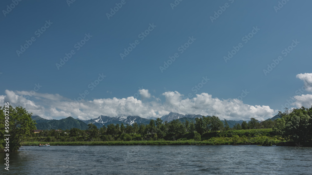 Fototapeta premium A blue calm river flows in the valley. A rafting boat with people is visible. Green vegetation on the shore. Picturesque mountains against the sky and clouds. Copy space. Kamchatka