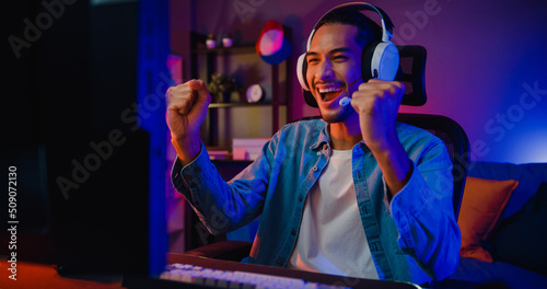 Happy asia guy gamer wear headphone participation play video game colorful neon lights computer in living room at night modern house. Esport streaming game online, Home quarantine activity concept. photo