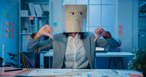 Young Asian businesswoman sit on desk wear draw sad mask paper craft bag feel bad mood with fail work project in office at night. Anonymous lady mad and confuse with overwork, Work mental health. photo