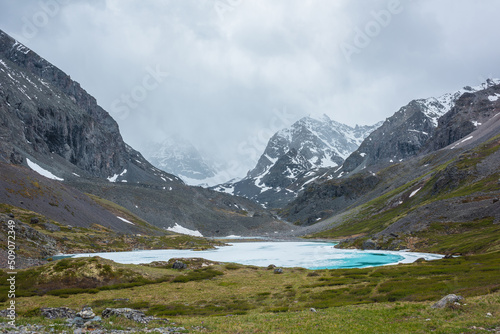 Atmospheric mountain landscape with frozen alpine lake and high snowy mountains. Awesome overcast scenery with icy mountain lake on background of snow mountains in low clouds. Scenic view to ice lake.