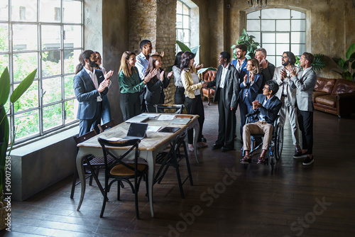 Two multi-ethnic business women sign an agreement with a handshake while colleagues clap together happy in office meeting room - concept of people, handshake agreement, promotion, signing a contract