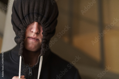 tense teenage guy in a black pulled-down hood covering the upper part of his face. Transitional age and adolescent problems. The teenager wants to hide from everyone.