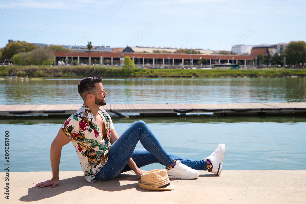 Handsome young man is wearing a shirt with Hawaiian flowers, a hat and is sitting on the river bank at the jetty sunbathing. Holiday and leisure time and travel concept.