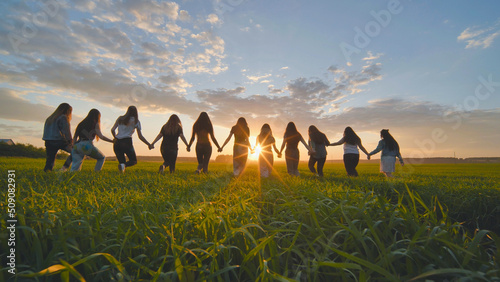 A group of girls walk towards the sun at sunset holding hands.