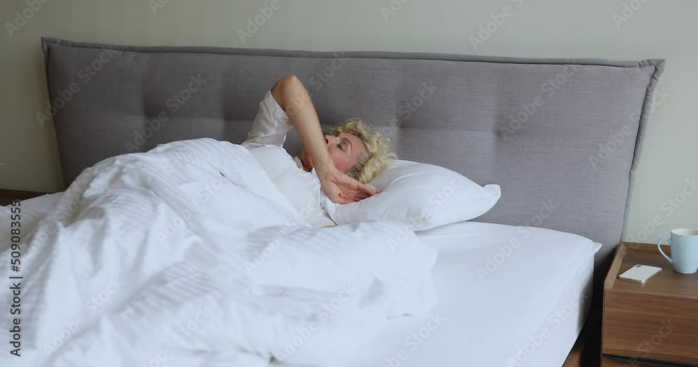 Attractive older 60s woman wake up looks rested and happy, sits down stretching spine raising up hands make morning exercise awakened in good mood feels refreshed after quality healthy sleep at home Attractive older 60s woman wake up looks rested and happy, sits down stretching spine raising up hands make morning exercise awakened in good mood feels refreshed after quality healthy sleep at home