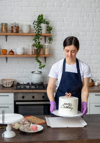 Pastry chef in an apron holds white cake with the inscription Happy birthday and numbers 32 in his hands and puts it on cardboard box. Selective focus. Photos about confectioners, food, hobbies.