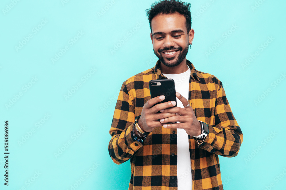 Handsome smiling hipster model. Sexy unshaven man dressed in yellow summer shirt and jeans clothes. Fashion male posing near blue wall. Holding smartphone. Looking at cellphone screen. Using apps Handsome smiling hipster model. Sexy unshaven man dressed in yellow summer shirt and jeans clothes. Fashion male posing near blue wall. Holding smartphone. Looking at cellphone screen. Using apps
