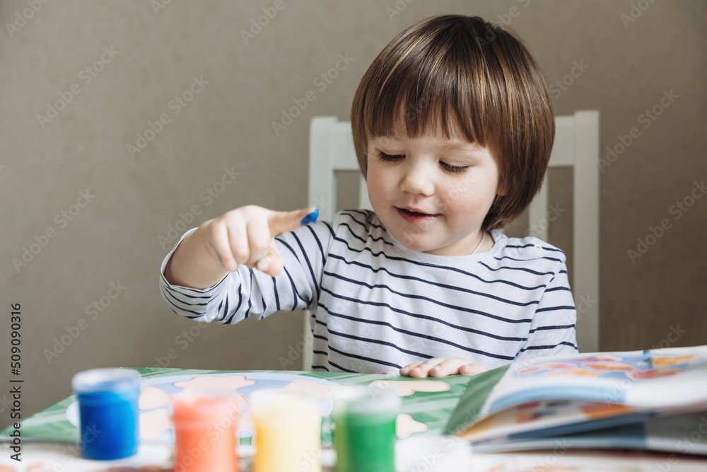 Finger painting. Portrait of cute little boy painting with fingers at home. Close-up of child's hand in colorful paints. Early education concept. Sensory play. Development of fine motor skills. Finger painting. Portrait of cute little boy painting with fingers at home. Close-up of child's hand in colorful paints. Early education concept. Sensory play. Development of fine motor skills.
