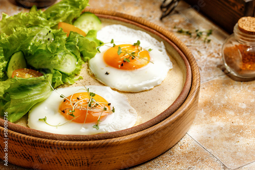 Fried eggs with micro greens, lettuce and cherry tomatoes on beige background, tile, in sunny light. Healthy breakfast in a wooden tray.