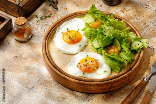 Two Fried eggs with lettuce and cherry tomatoes on beige background, tile, in sunny light. Healthy breakfast is served in a wooden tray.