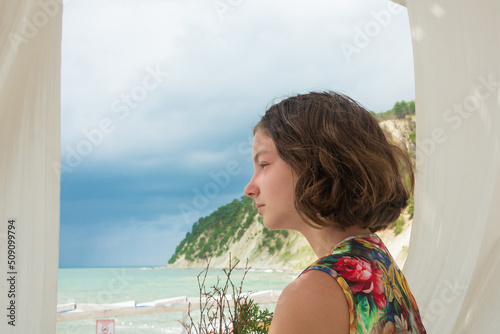 Teenage girl looks at the sea on a summer terrace with white curtains. Side view in profile