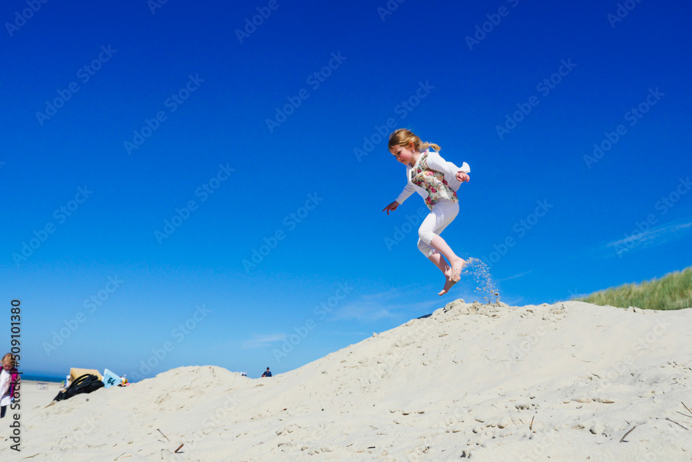 Little girl jumping on sand dune with blue sky background. Profile view. Long shot.