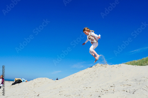 Little girl jumping on sand dune with blue sky background. Profile view. Long shot.