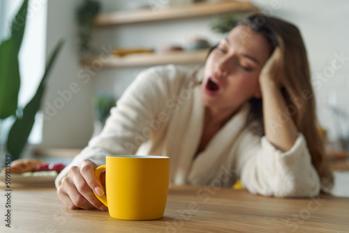 Young woman in bathrobe yawning with cup of coffee while sitting at the kitchen counter at home photo