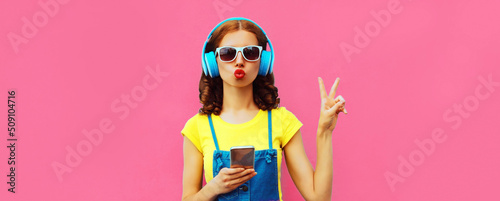 Portrait of stylish young woman in headphones listening to music with smartphone on pink background
