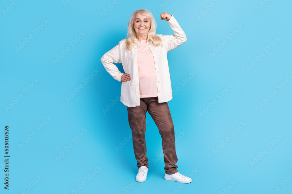 Full size photo of cheerful mature woman show hand muscles healthy sportive isolated over blue color background Full size photo of cheerful mature woman show hand muscles healthy sportive isolated over blue color background