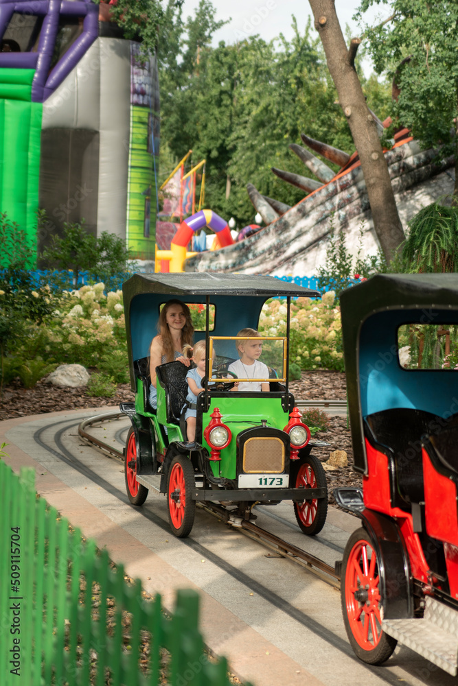 Young mother with two children in Amusement park. Family ride in small toy car. Nanny rides with children in the park.