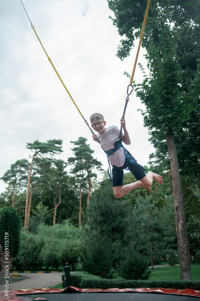 Boy hangs on slings, jumps high on trampoline in an amusement park. Weekend at theme park. Teenager is having fun.