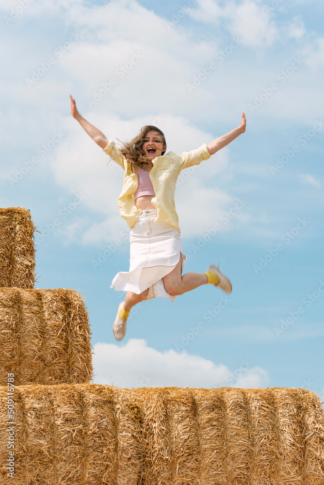 Cheerful girl jumps from high haystack. Happy young woman has fun on the farm on blue sky background. Cheerful girl jumps from high haystack. Happy young woman has fun on the farm on blue sky background.
