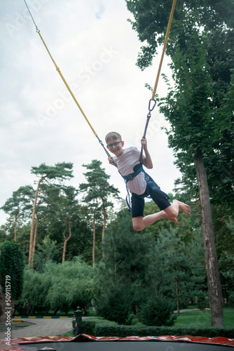 Boy hangs on slings, jumps high on trampoline in an amusement park. Weekend at theme park. Teenager is having fun.