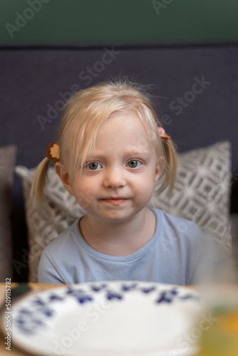 Portrait of sad little blonde girl with two tails in cafe. Hungry three-year-old girl sitting with empty plate at the table. Waiting for meal