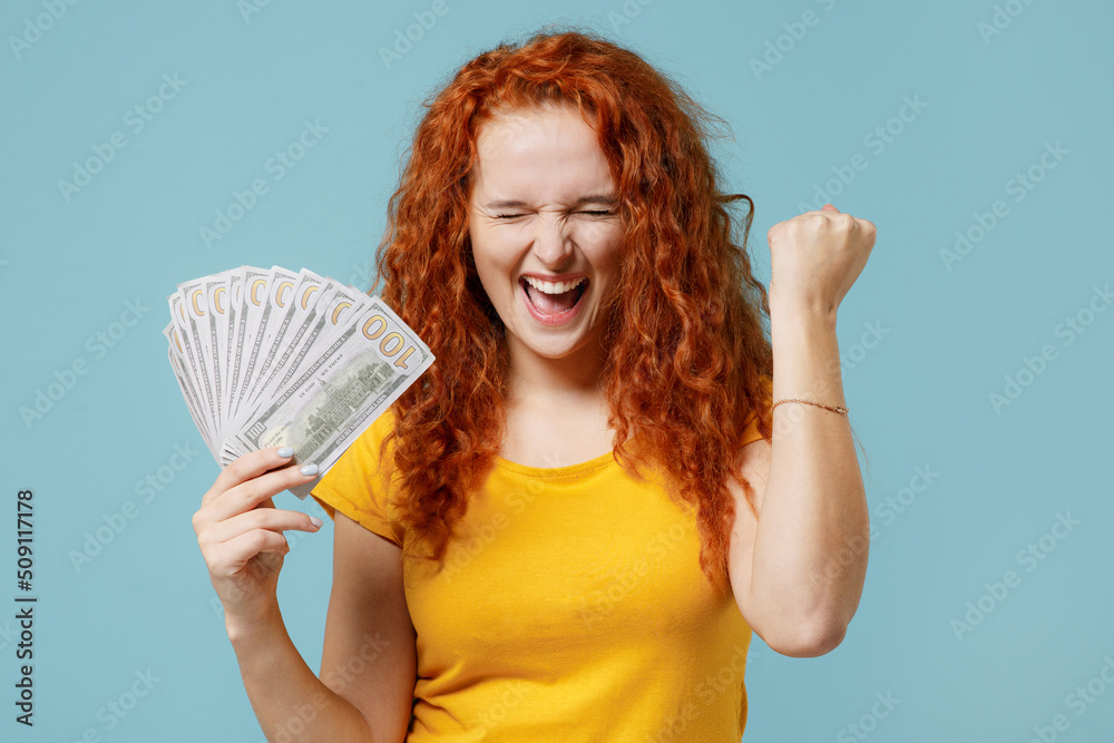Young fun happy redhead woman 20s wear yellow t-shirt holding fan of cash money in dollar banknotes do winner gesture isolated on plain light pastel blue background studio. People lifestyle concept Young fun happy redhead woman 20s wear yellow t-shirt holding fan of cash money in dollar banknotes do winner gesture isolated on plain light pastel blue background studio. People lifestyle concept