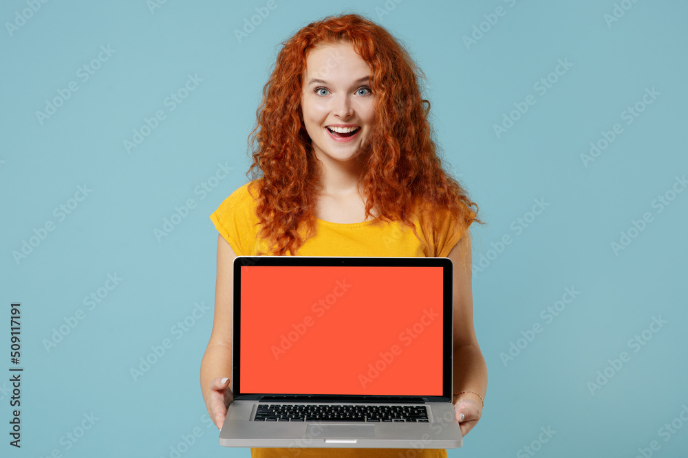 Young surprised fun happy redhead woman 20s wearing yellow t-shirt hold use work on laptop pc computer with blank screen workspace area isolated on plain light pastel blue background studio portrait Young surprised fun happy redhead woman 20s wearing yellow t-shirt hold use work on laptop pc computer with blank screen workspace area isolated on plain light pastel blue background studio portrait