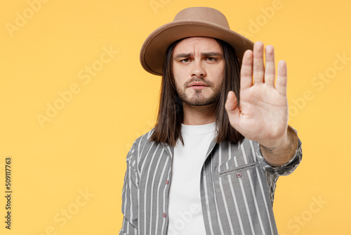 Young strict serious sad man he 20s wears striped grey shirt white t-shirt hat showing hand stop gesture with palm isolated on plain yellow color background studio portrait. People lifestyle concept.