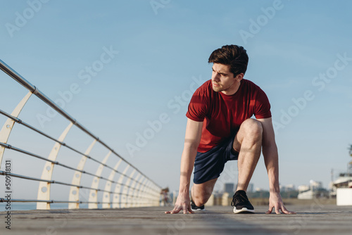 Full length young strong sporty toned fit sportsman man 20s wear sports clothes stand at low start going to run warm up training at sunrise sun dawn over sea beach outdoor on pier seaside in morning © ViDi Studio