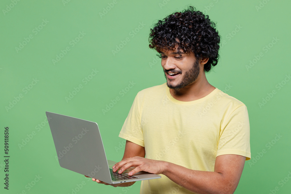Young smiling happy Indian man 20s in basic yellow t-shirt hold use work on laptop pc computer chatting online isolated on plain pastel light green background studio portrait People lifestyle concept - obrazy, fototapety, plakaty 