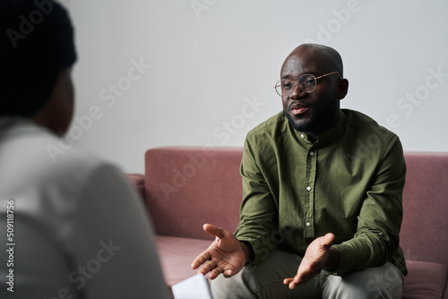 Young black man in casualwear sitting on couch and explaining his emotions and worries during talk with psychoanalyst