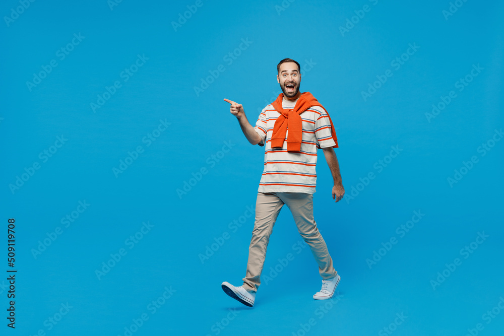 Full body side profile view young amazed fun man 20s wearing orange striped t-shirt walking going point index finger aside isolated on plain blue background studio portrait. People lifestyle concept. Full body side profile view young amazed fun man 20s wearing orange striped t-shirt walking going point index finger aside isolated on plain blue background studio portrait. People lifestyle concept.