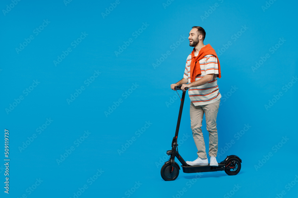 Full body side view young smiling happy caucasian man 20s wearing orange striped t-shirt riding electric scooter walking isolated on plain blue background studio portrait. People lifestyle concept Full body side view young smiling happy caucasian man 20s wearing orange striped t-shirt riding electric scooter walking isolated on plain blue background studio portrait. People lifestyle concept