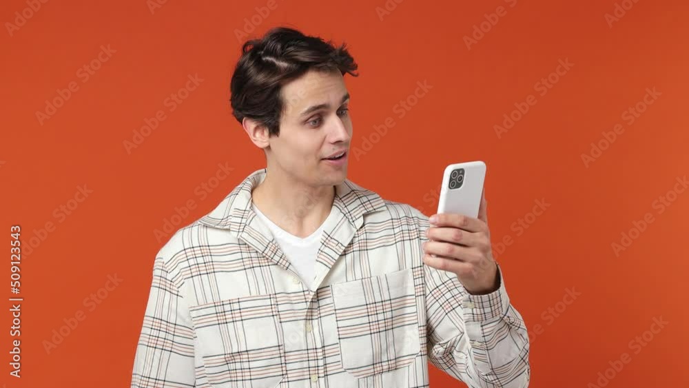 Young brunet man 20s wears white shirt get video call using mobile cell phone do selfie talk conducting pleasant conversation greet with hand isolated on plain orange wall background studio portrait Young brunet man 20s wears white shirt get video call using mobile cell phone do selfie talk conducting pleasant conversation greet with hand isolated on plain orange wall background studio portrait