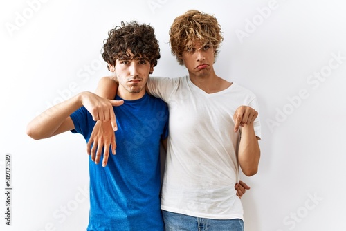 Young gay couple standing together over isolated background pointing down looking sad and upset, indicating direction with fingers, unhappy and depressed.