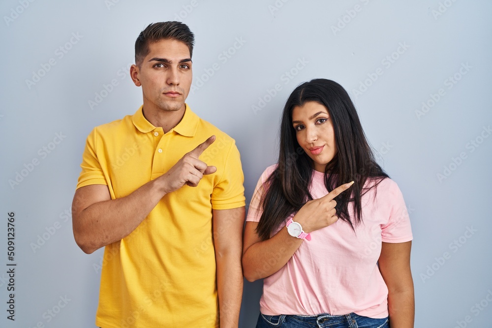 Young couple standing over isolated background pointing with hand finger to the side showing advertisement, serious and calm face Young couple standing over isolated background pointing with hand finger to the side showing advertisement, serious and calm face