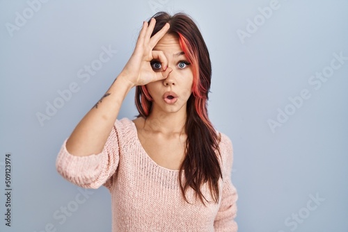 Young caucasian woman wearing pink sweater over isolated background doing ok gesture shocked with surprised face, eye looking through fingers. unbelieving expression.