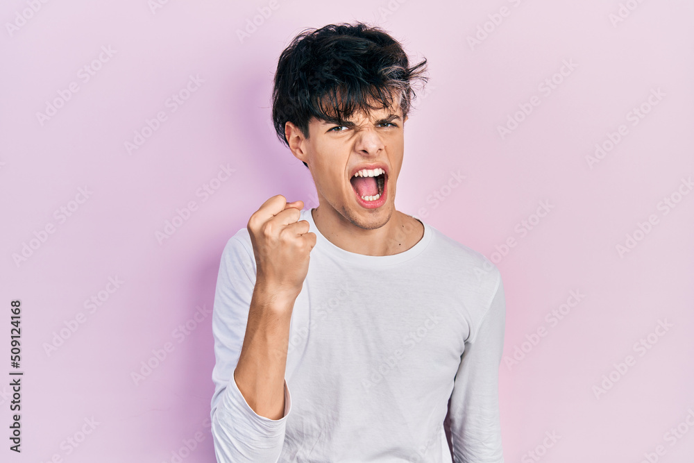 Handsome hipster young man wearing casual white shirt angry and mad raising fist frustrated and furious while shouting with anger. rage and aggressive concept. Handsome hipster young man wearing casual white shirt angry and mad raising fist frustrated and furious while shouting with anger. rage and aggressive concept.