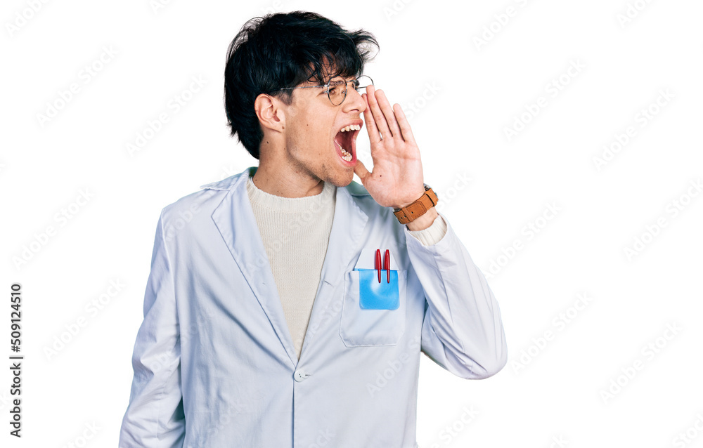 Handsome hipster young man wearing doctor uniform shouting and screaming loud to side with hand on mouth. communication concept. Handsome hipster young man wearing doctor uniform shouting and screaming loud to side with hand on mouth. communication concept.