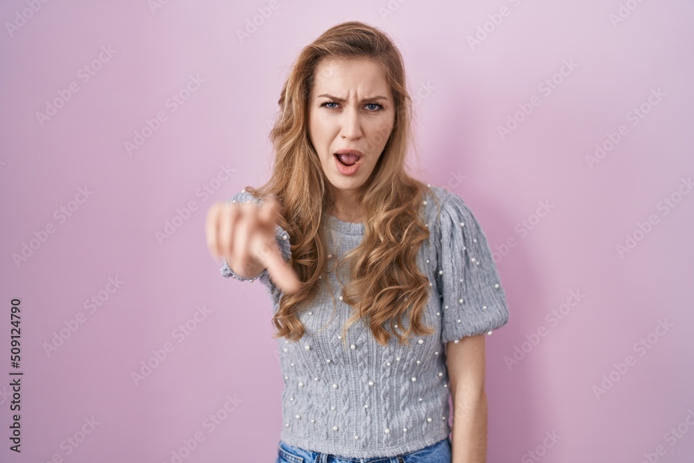 Beautiful blonde woman standing over pink background pointing displeased and frustrated to the camera, angry and furious with you Beautiful blonde woman standing over pink background pointing displeased and frustrated to the camera, angry and furious with you