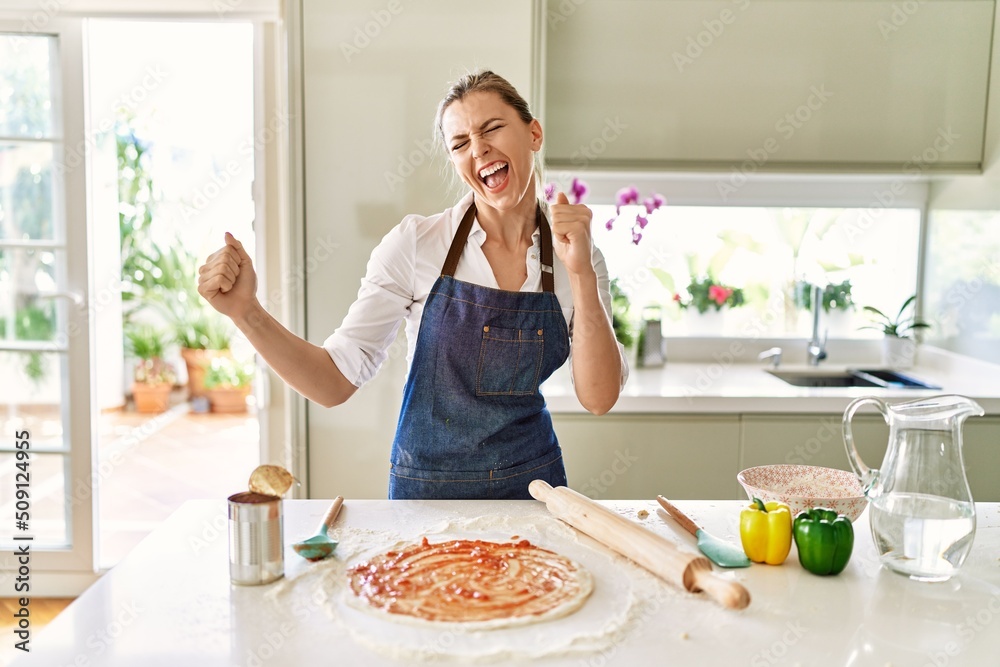 Beautiful blonde woman wearing apron cooking pizza very happy and excited doing winner gesture with arms raised, smiling and screaming for success. celebration concept.