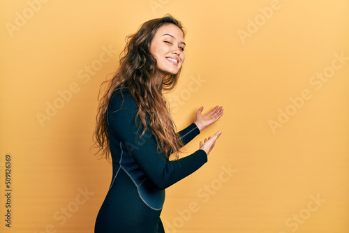 Young hispanic girl wearing diver neoprene uniform inviting to enter smiling natural with open hand